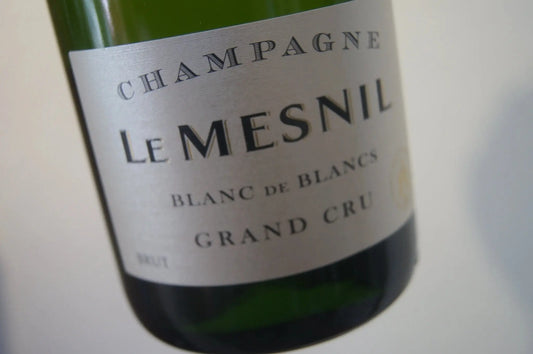 Eine Flasche Le Mesnil Grand Cru Blanc de Blancs Champagner, umgeben von perfekten Paarungen wie Fisch, Ziegenkäse und frischem Gemüse. Erleben Sie die Eleganz und den luxuriösen Geschmack, unterstützt durch die einzigartige Kreidebodenstruktur von Mesnil, die den Chardonnay-Trauben ihre außergewöhnliche Mineralität und Tiefe verleiht.