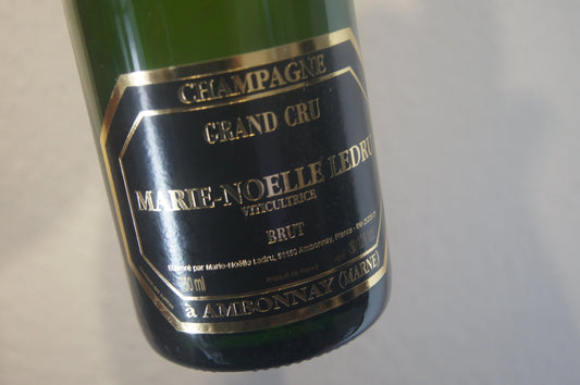 Die Flasche Marie-Noelle Ledru Champagner ruht in einem Weinkeller, umgeben von der dunklen Eleganz von Eichenfässern, bereit, die tiefe Mineralität und reiche Frucht des Pinot Noir aus Ambonnay zu enthüllen. Ein Symbol für unvergleichliche Qualität.